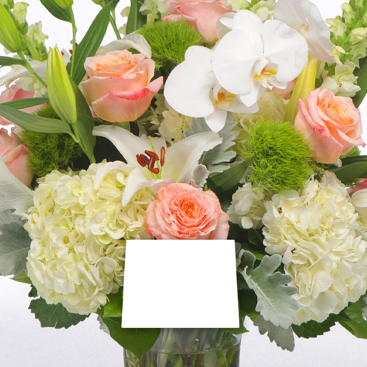 A bouquet sits in a glass vase, featuring delicate light pink roses, white lilies, and hydrangeas, accented by green foliage. A blank white card is attached to the arrangement.