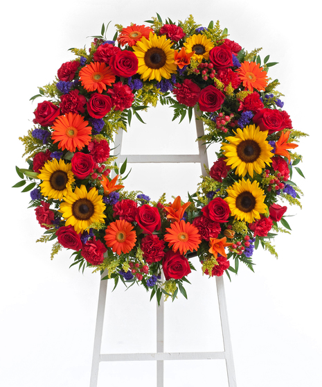 A wreath arranged with roses, carnations, sunflowers, lilies, gerbera daisies, mixed with greenery and other seasonal flowers. Displayed against a white background.