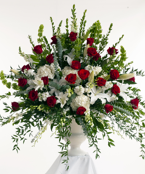 A large, lush floral arrangement with red roses, white lilies, hydrangeas, and snapdragons, accented by green leaves, displayed in a white urn against a white background.