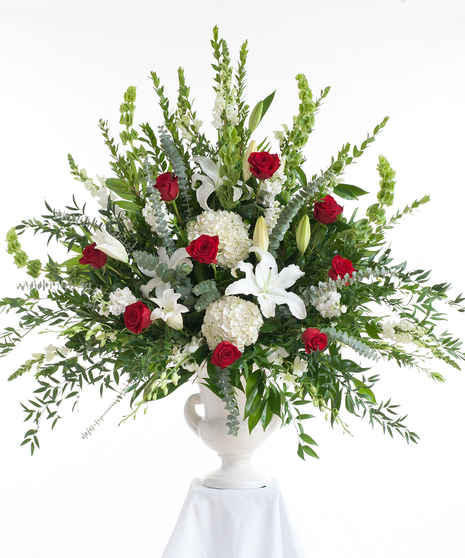 A large, lush floral arrangement with red roses, white lilies, hydrangeas, and snapdragons, accented by green leaves, displayed in a white urn against a white background.