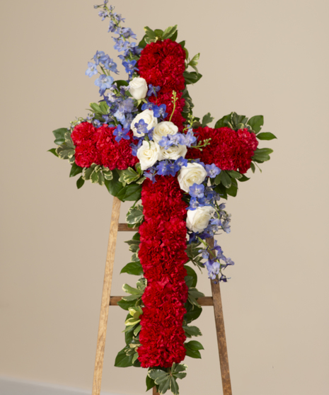 A cross-shaped floral arrangement made of red carnations adorned with white roses, blue delphinium and greenery, displayed on an easel.