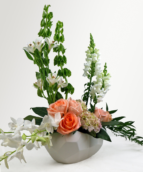 Image showcases a floral blend of peach hydrangea, coral roses, dendrobiums, bells of Ireland, alstroemeria, and snapdragons in a geometric long white ceramic container. 