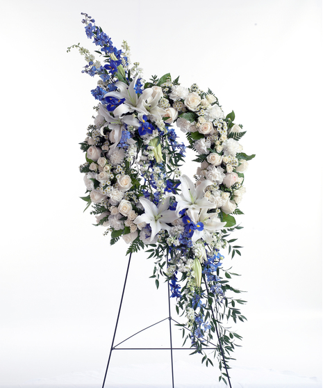 Circular floral wreath made of white lilies, roses, and carnations, accented by blue delphinium and greenery, displayed on a stand against a white background.