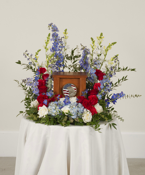 A patriotic arrangement of roses, hydrangeas, carnations, and delphinium, mixed with greenery surrounding the urn. Displayed on a white table.