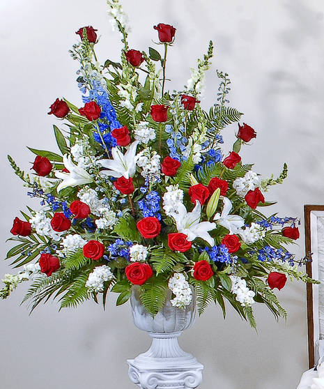 A large, lush floral arrangement with red roses, white lilies, blue delphinium, and stock, accented by green leaves. Displayed in a white urn.