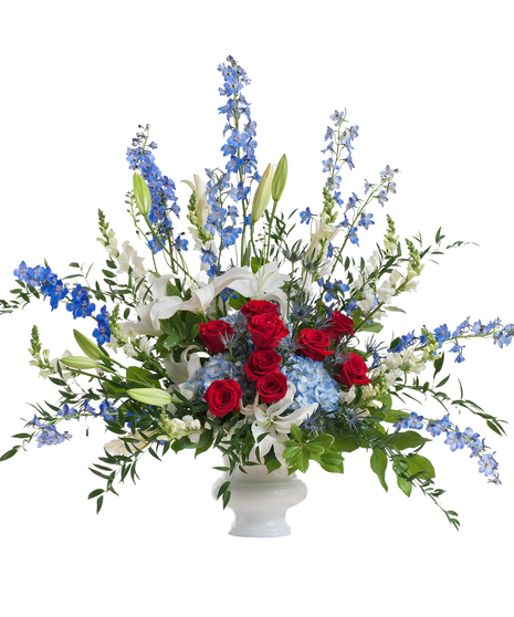 A design of roses, lilies, hydrangeas, and delphinium, mixed with greenery. Placed in a white urn, displayed against a white background.