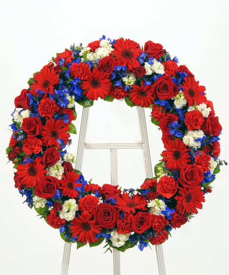 A circular wreath of red roses, gerbera daisies, carnations, and stock. Displayed on a white easel against a white background.