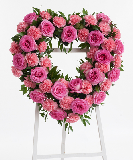 A heart-shaped wreath of roses and carnations, mixed with greenery. Displayed on a white easel against a white background.