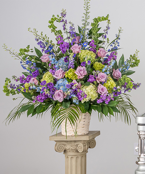 An arrangement of roses, hydrangeas, delphinium, bells of Ireland, and other seasonal flowers in a white basket.