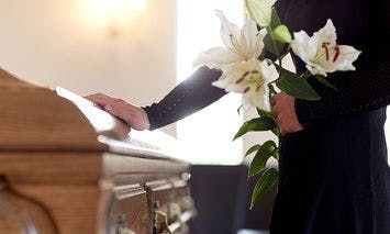 A person in black attire stands by a wooden casket, holding white lilies in a well-lit room with soft, warm lighting.